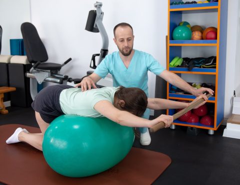 Kinetoterapeut asista o pacienta in timpul unui exercitiu de gimnastica medicala realizat pe o minge de fitness , pentru corectarea posturii si tonifiere musculara